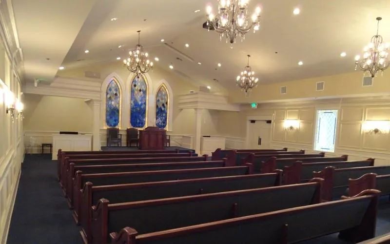 A funeral home chapel with rows of wooden pews, chandeliers, and tall stained-glass windows in soft light